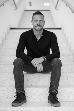 Auf dem schwarz-weiß Bild ist ein Mann abgebildet, der auf einer Treppe sitzt und in die Kamera lächelt.